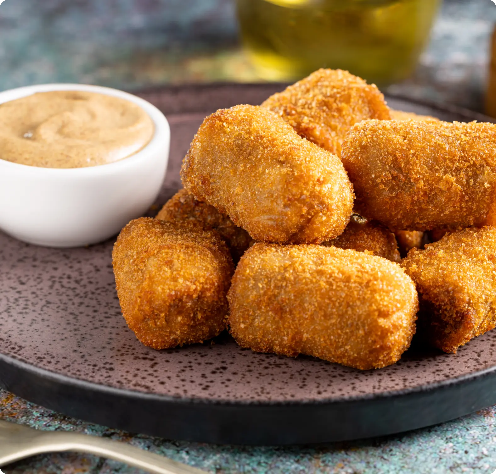 A plate of crispy, golden-brown croquettes with a small bowl of creamy dipping sauce on the side.