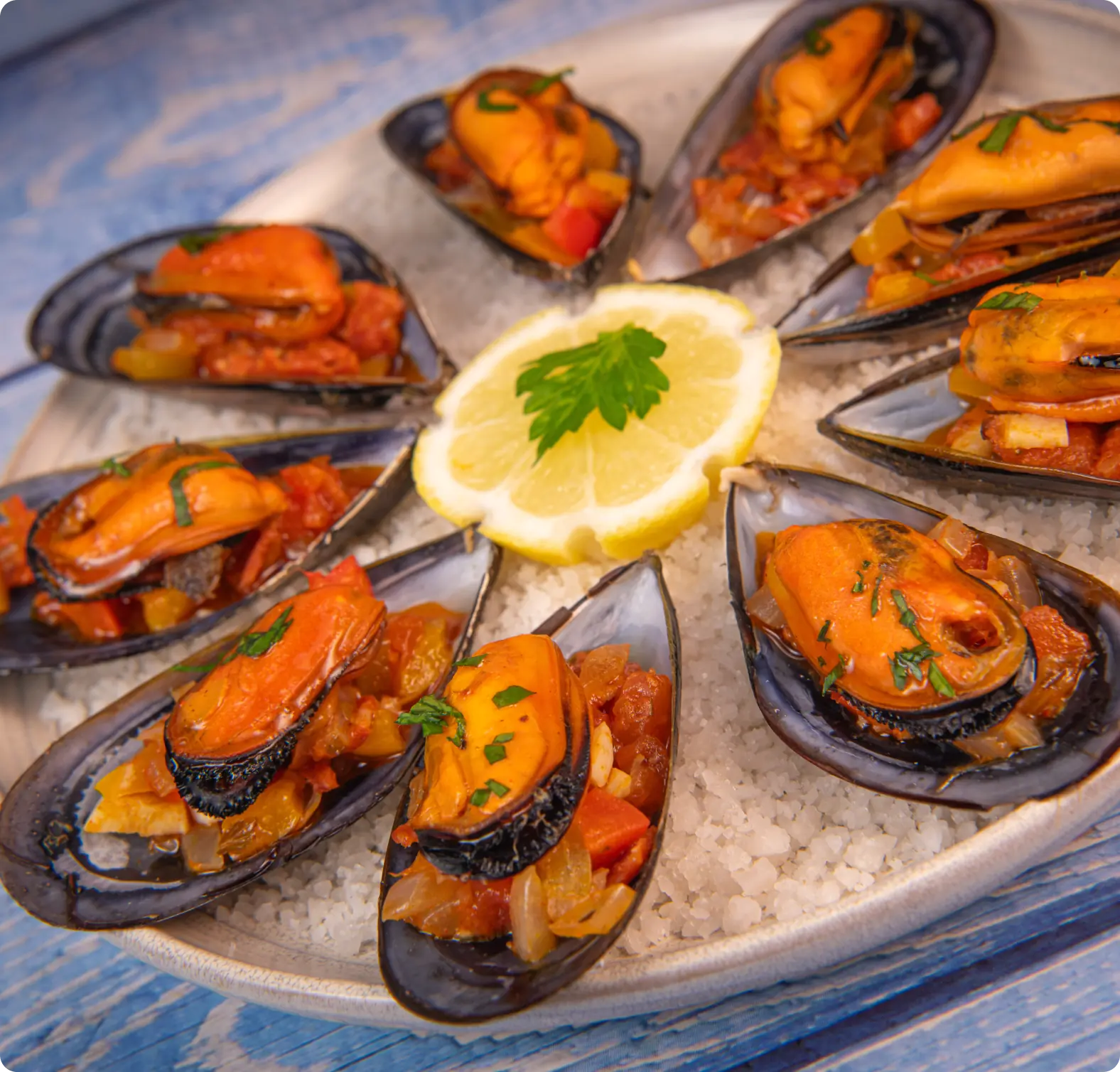 A plate of mussels served in shells with a tomato-based sauce on a bed of salt, garnished with a lemon slice and parsley.