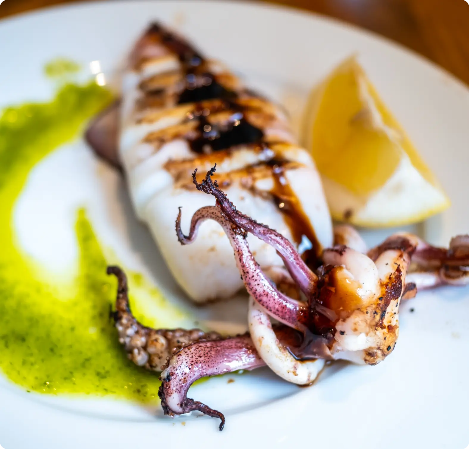 Grilled squid with tentacles, drizzled with dark sauce, served on a white plate with a wedge of lemon and a swirl of green sauce.